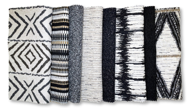 An array of fabric swatches showcasing a variety of textures and patterns in black, white, and gray tones. From left to right: a bold geometric pattern, a textured solid gray, a narrow stripe, a speckled tweed, a darker solid, a high-contrast ikat design, and a fabric featuring a tribal-inspired print with gold accents.