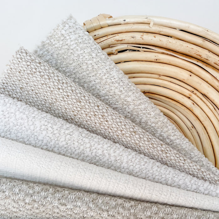 A collection of textured upholstery fabrics in varying shades of white and light gray, neatly folded and stacked in ascending order of texture complexity, displayed against a natural, curved rattan chair back.