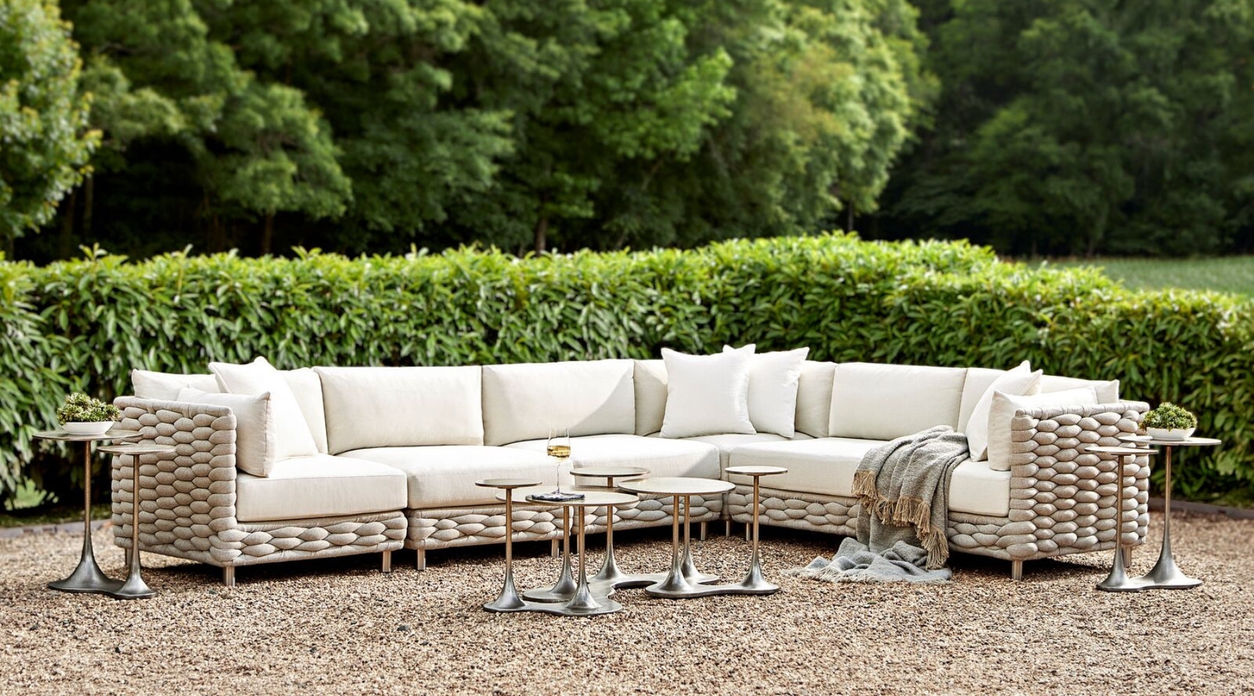 Outdoor luxury seating area with a large L-shaped off-white sectional sofa, adorned with matching cushions. The sofa features unique pebble-like detailing on the sides. Set on a pebbled patio, the setup includes sleek, modern metal side tables of varying heights, with a glass of white wine on one and small potted plants on others. The area is framed by a lush green hedge, emphasizing the serene and manicured garden ambiance.