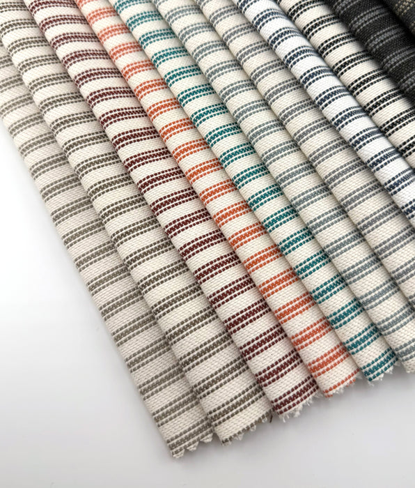 Assorted Revolution Performance Upholstery Fabric swatches from the Foreshore collection arranged in a fanned layout, showing multiple colorways including sand, walnut, conch, mint, bottle, mango, red, marine, onyx, and carbon. Each swatch displays Foreshore’s evenly spaced vertical stripe pattern with subtle tonal variation.