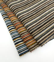 Assorted Revolution Performance Upholstery Fabric swatches arranged in a fanned layout. Each swatch displays Mitchell’s narrow vertical stripe pattern with a tightly woven, subtly raised texture and soft zigzag edges, creating layered tonal variation across the surface.