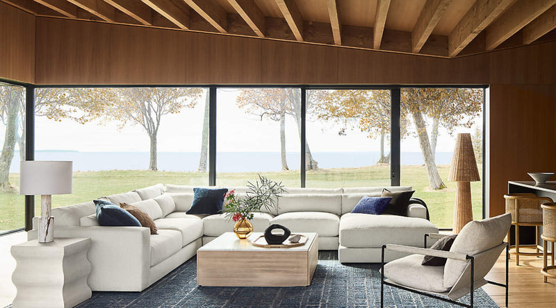 Here is the alt-text for the image you provided:

"A modern living room with a large L-shaped white sectional sofa, accented with blue and brown pillows, set against floor-to-ceiling windows offering a view of a serene lakeside landscape. The room features a wooden ceiling with exposed beams, a light wooden coffee table, and a decorative lamp on a unique wavy-shaped side table. A sleek armchair and woven barstools complete the minimalist yet cozy design, with natural light streaming in and highlighting the 