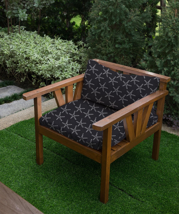 Lifestyle image of Revolution Performance Upholstery Fabric, pattern Seastar in Onyx, shown upholstered on a wooden outdoor lounge chair. The woven starfish motif appears in soft ivory against a deep charcoal ground, creating a lightly textured surface with crisp definition and a bold, graphic coastal look.