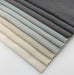 Assorted Revolution Performance Upholstery Fabric swatches from the Shelter collection arranged in a fanned layout, showing multiple colors including ivory, cream, beige, stone, light grey, silver grey, and charcoal variations. Each swatch displays Shelter’s smooth, tightly woven texture with subtle tonal variation.
