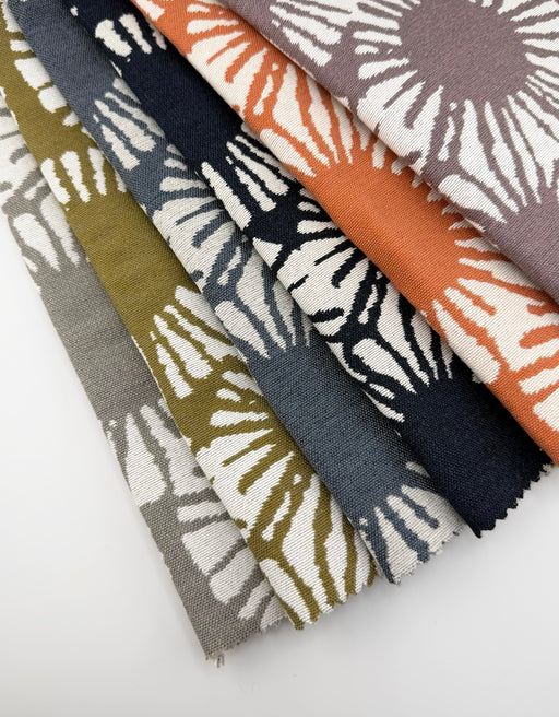 Revolution Performance Fabric Sunburst pattern swatches arranged in a fanned layout, showcasing multiple colorways including citron, indigo, mango, lapis, loft, and passion, each featuring bold radiating sunburst motifs on an ivory ground with a textured, high-performance upholstery weave.